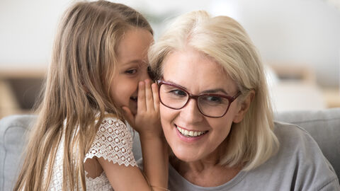 Kleines Mädchen lehnt sich zu Großmutter herüber und flüstert ihr ins Ohr, diese lächelt und hört gespannt zu.