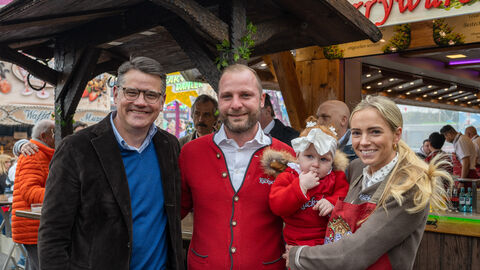Ministerpräsident Rhein neben einem Mann und einer Frau, die ein kleines Kind auf dem Arm trägt, vor einer Bude auf der Dippemess