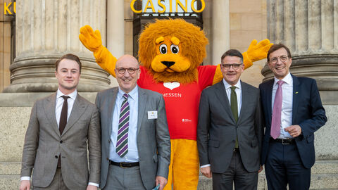 Chef der Staatskanzlei, Staatssekretär Benedikt Kuhn, VhU-Hauptgeschäftsführer Dirk Pollert, Ministerpräsident Boris Rhein, VU-Präsident Wolf Matthias Mang Vier Männer stehen stehen vor einem Gebäude. In der Mitte steht eine Person als plüschiger Löwe angezogen.