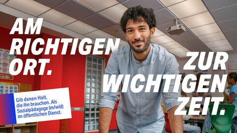 Ein Sozialpädagoge steht im Klassenzimmer. Auf dem Bild steht der Text: "Am richtigen Ort zur wichtigen Zeit. Gib denen Halt, die ihn brauchen. Als Sozialpädagoge (m/w/d) im öffentlichen Dienst."