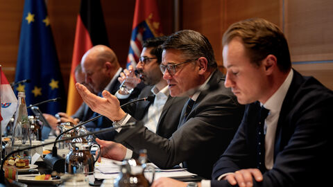 Politik und Wirtschaft sprechen beim Rüstungsgipfel über die Stärkung der heimischen Verteidigungs- und Sicherheitsindustrie. Ministerpräsident Rhein, Staatssekretär Kuhn und weitere Mitglieder der Landesregierung sitzen am Tisch während der Gespräche des Regierungsgipfels. Ministerpräsident Rhein spricht in ein Mikrofon.