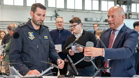 Ein Mann in Polizeiuniform und ein Mann in Anzug stehen vor einem Tisch und halten eine Drone hoch.